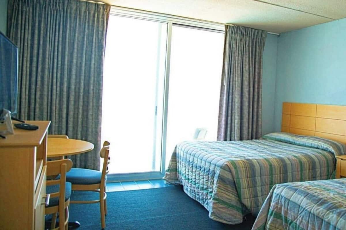 A hotel room with a desk, two beds, a chair, a lamp, and a window with curtains.