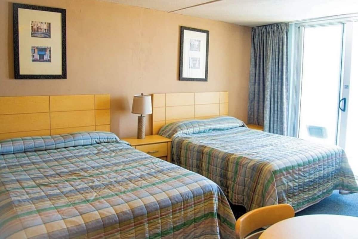 A hotel room with two beds, a nightstand with a lamp, a table, and a sliding door to the outside.