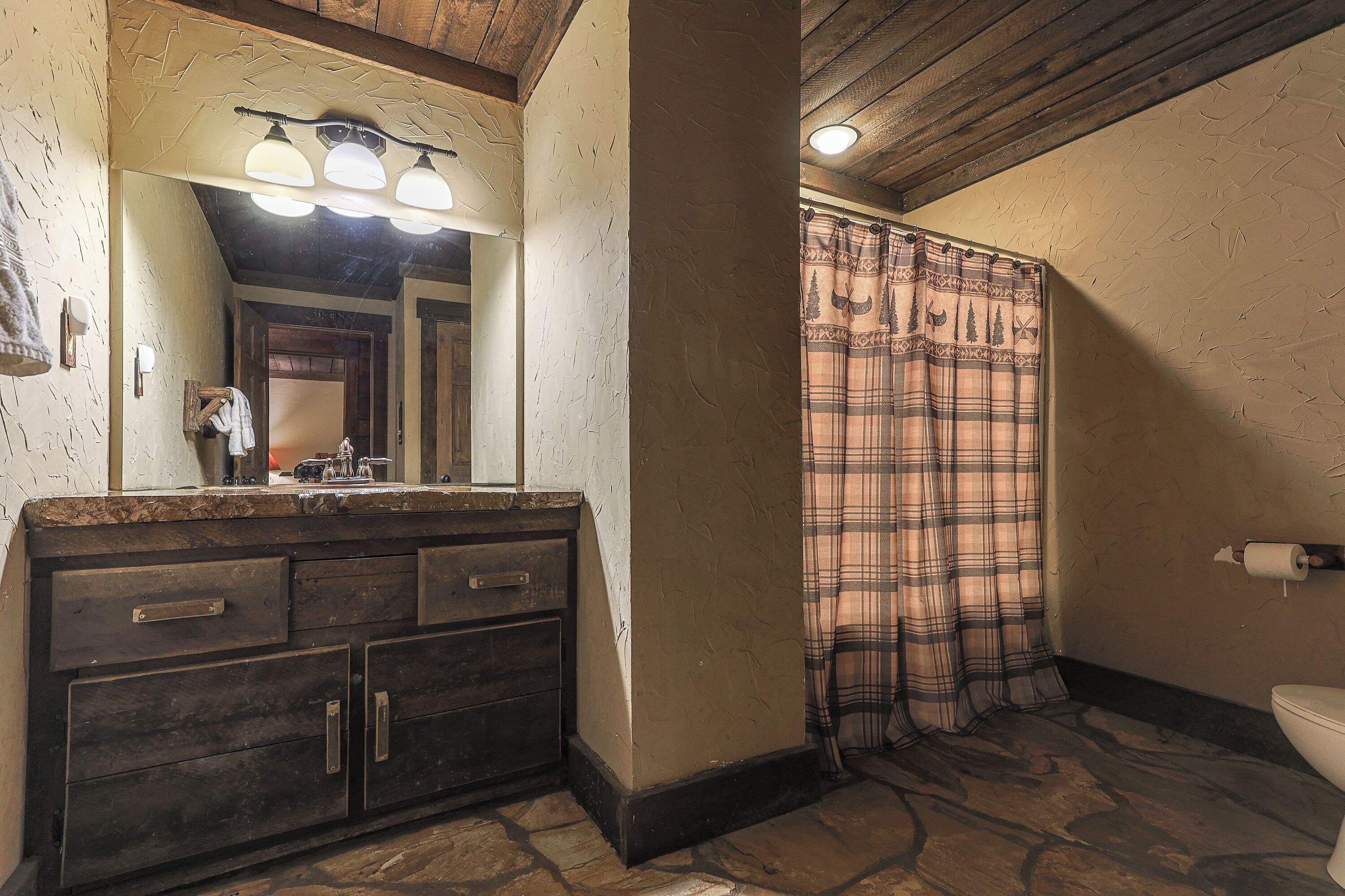 A bathroom with a stone floor, a wooden vanity, a large mirror, and a plaid shower curtain.
