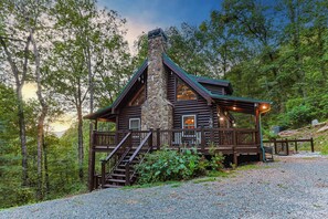 A log cabin with a stone chimney, wooden deck, and a gravel driveway.