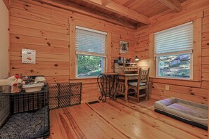 A cozy cabin interior with wooden walls, a dining table, a pet bed, and a window with a view of the outdoors.