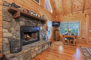 A log cabin interior with a stone fireplace, wooden shelves, a television, and a dining area.