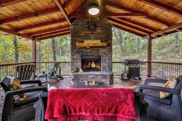 Una terraza techada con chimenea de piedra, mobiliario exterior y vista al bosque.