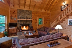 A cozy cabin interior with a stone fireplace, wooden walls, a sofa, a television, and a staircase. - Hidden Valley Paradise | Ellijay, GA (Ellijay)