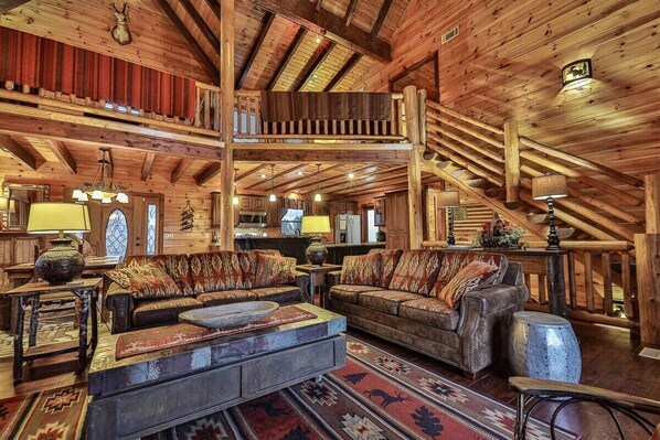 A log cabin interior with a loft bedroom, rustic furniture, and a chandelier.