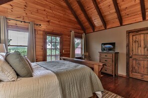 A wooden cabin bedroom with a bed, a dresser, a television, and a window with curtains. - Fox Run Ridge | Ellijay, GA (Ellijay)