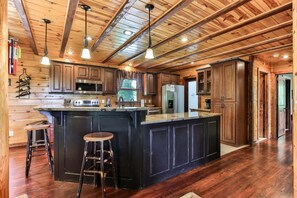 A spacious kitchen with wooden cabinetry, a central island with stools, stainless steel appliances, and a high ceiling with exposed beams. - Fox Run Ridge | Ellijay, GA (Ellijay)