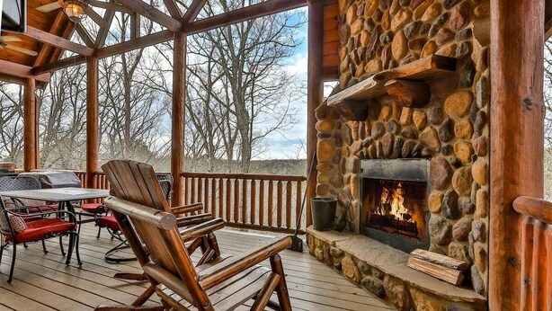 A wooden deck with a stone fireplace, outdoor seating, and a view of trees.