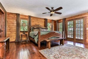 A wooden cabin bedroom with a bed, a nightstand, a lamp, a ceiling fan, and large windows. - Fox Run Ridge | Ellijay, GA (Ellijay)