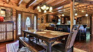 A rustic wooden interior with a dining table set for four, wooden beams on the ceiling, and a kitchen area in the background.