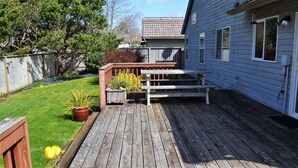 Un terrazzo in legno con una panchina e un tavolo, circondato da un giardino con piante e un prato.