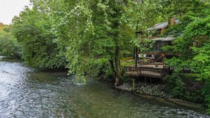 A riverside wooden structure with a deck, surrounded by dense greenery. - Lofty Pines on the Cartecay | Ellijay, GA (Ellijay)