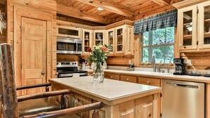 A wooden cabin interior with a kitchen featuring wooden cabinets, a central island, a dining table, and a window with a view of greenery. - Lofty Pines on the Cartecay | Ellijay, GA (Ellijay)