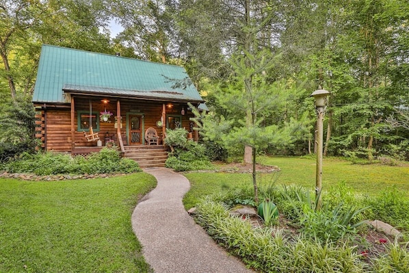 A rustic log cabin with a green roof, a porch with a bench, and a garden path leading to the entrance.