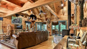 A log cabin interior with a wooden ceiling, a brown leather sofa, a glass coffee table, and a fireplace. - Lofty Pines on the Cartecay | Ellijay, GA (Ellijay)