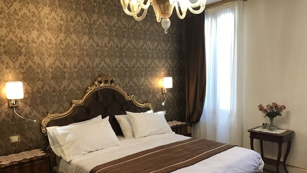 Una habitación de hotel con una cama grande, dos mesitas de noche, una lámpara auxiliar, un candelabro, un jarrón con flores y una ventana con cortinas.