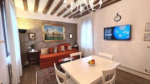 Un espacio de sala acogedor con un sofá, una mesa de comedor con sillas, una araña, un cuadro y un televisor en la pared.