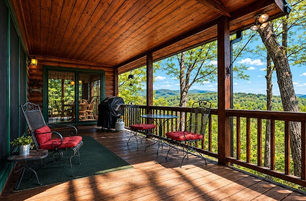 A covered deck with outdoor seating, a grill, and a view of a forested landscape.