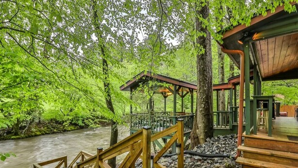Une terrasse en bois dotée d’une balustrade, qui offre une vue sur une rivière et est entourée d’un feuillage verdoyant et luxuriant.