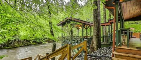 A wooden deck with a railing overlooking a river, surrounded by dense green foliage.