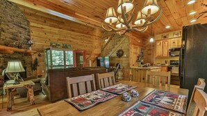 A rustic cabin interior with a wooden dining table, chairs, a chandelier, a fireplace, and a kitchen area. - Whitetail Ridge | Ellijay, GA (Ellijay)