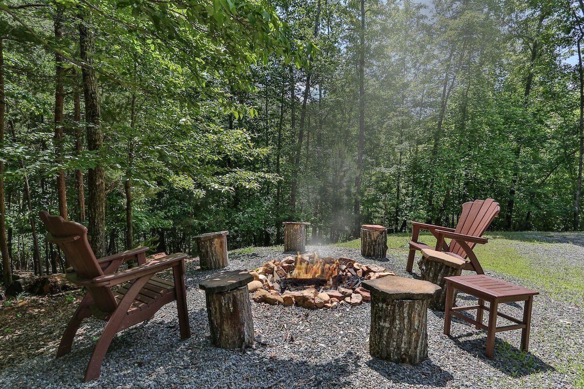 A forest setting with a fire pit surrounded by Adirondack chairs.