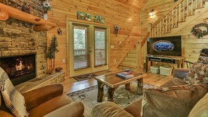 A cozy cabin interior with a fireplace, wooden walls, a television, and a staircase. - Whitetail Ridge | Ellijay, GA (Ellijay)
