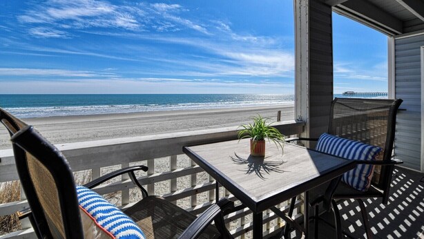 A balcony with a view of the beach and ocean, a table with a plant, and two chairs with striped cushions. - Sea Watch VIII 102 - Two Bedroom, Two Bath Oceanfront Condo (Garden City Beach)