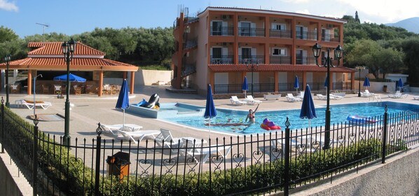 A pool area with lounge chairs and umbrellas, a covered seating area, and a building with balconies. - Zante Dolphin (Zakynthos)