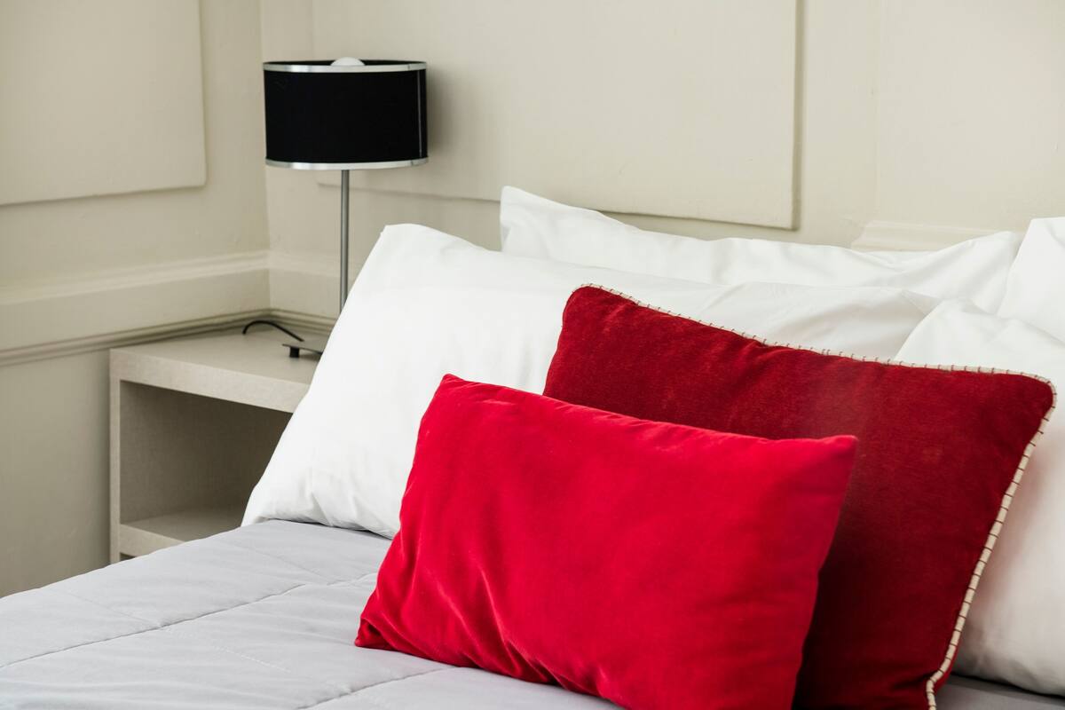 A neatly made bed with white and red pillows, a grey bedspread, and a black lamp on a bedside table.