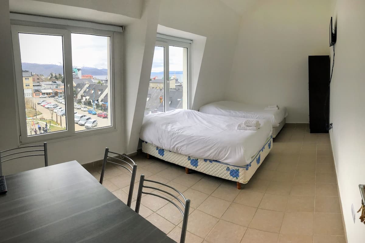 A small, clean room with two beds, a dining table, and a view of a cityscape through large windows.