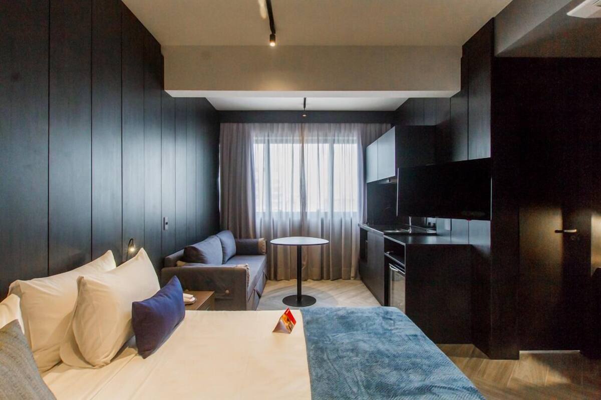 A modern hotel room with a large bed, a sofa, a small round table, a TV, and a kitchenette.