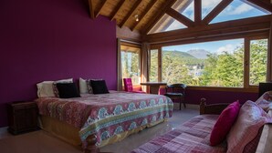 A bedroom with a large bed, a wooden headboard, a small table, and a view of a mountainous landscape through large windows.