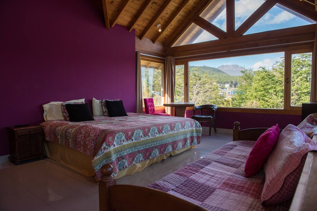 Une chambre à coucher avec un grand lit, une tête de lit en bois, une petite table et une vue sur un paysage montagneux grâce à de grandes fenêtres.
