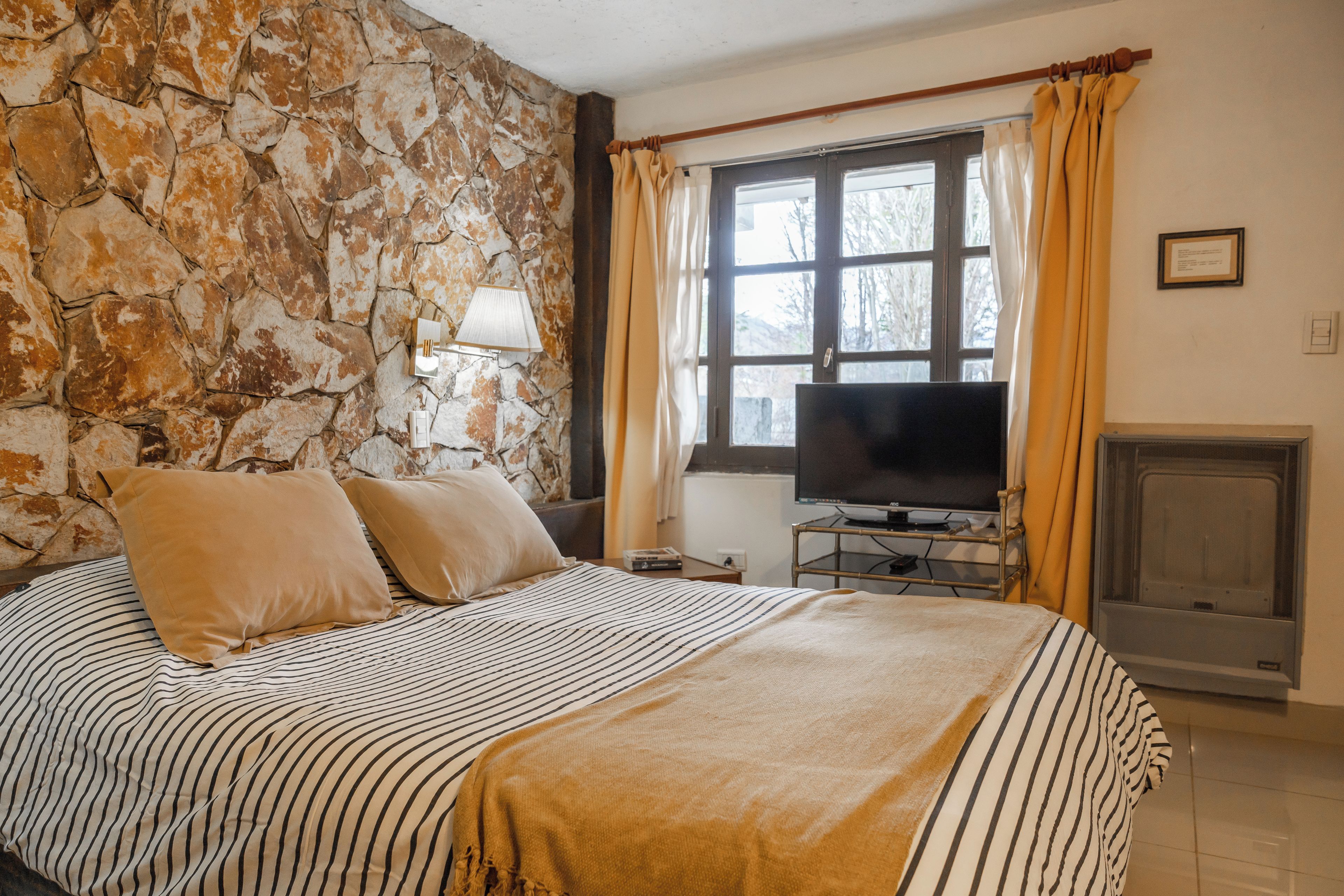 A bedroom with a stone wall, a bed with striped bedding, a television, and a fireplace.
