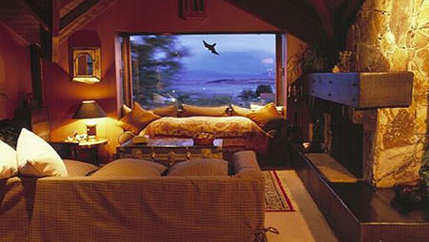A cozy room with a bed, a window with a view of the outdoors, a lamp, and a fireplace.