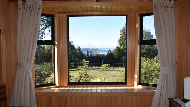 A bedroom with a large window offering a view of a grassy area and trees. - Vista Tranquila Chiloe (Chonchi)