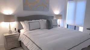 A neatly made bed with white bedding and black trim, flanked by two bedside tables with lamps and a phone. A large abstract painting hangs above the bed. - One Bedroom Suite (Boston)