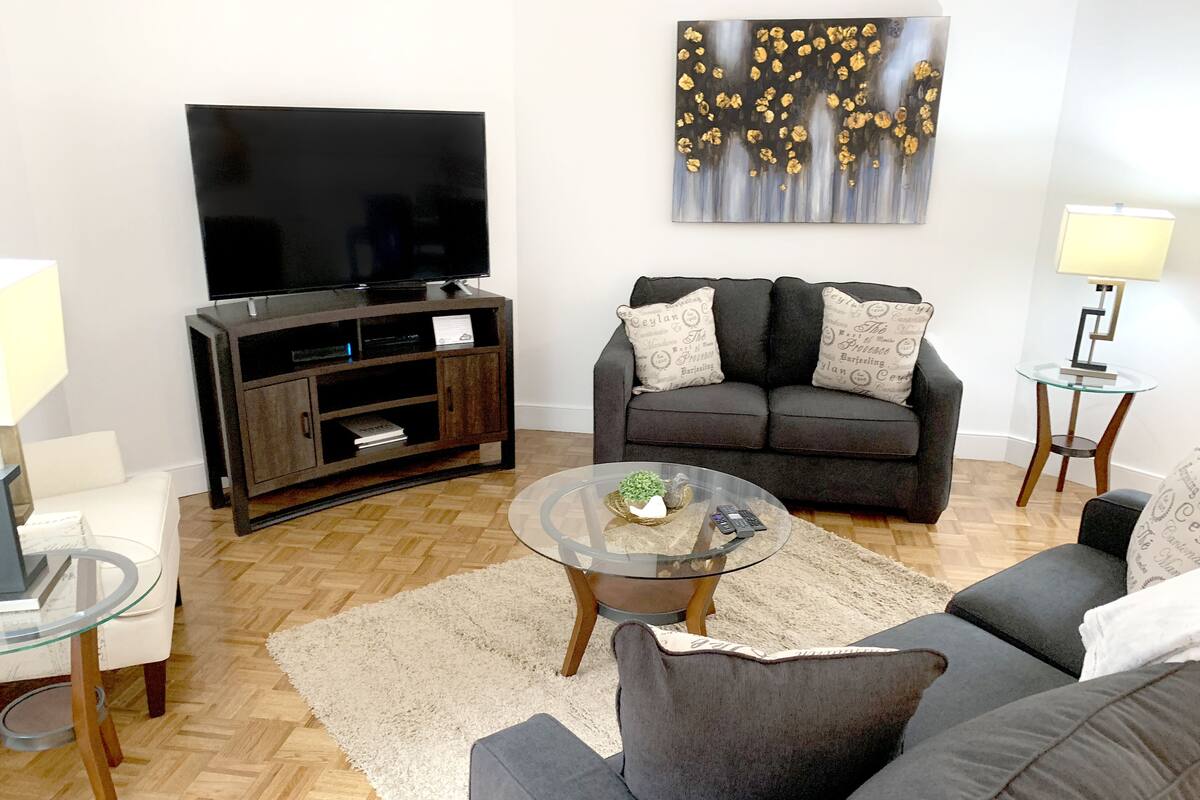 A living room with a flat-screen TV, dark sofas, a glass coffee table, and a decorative painting.