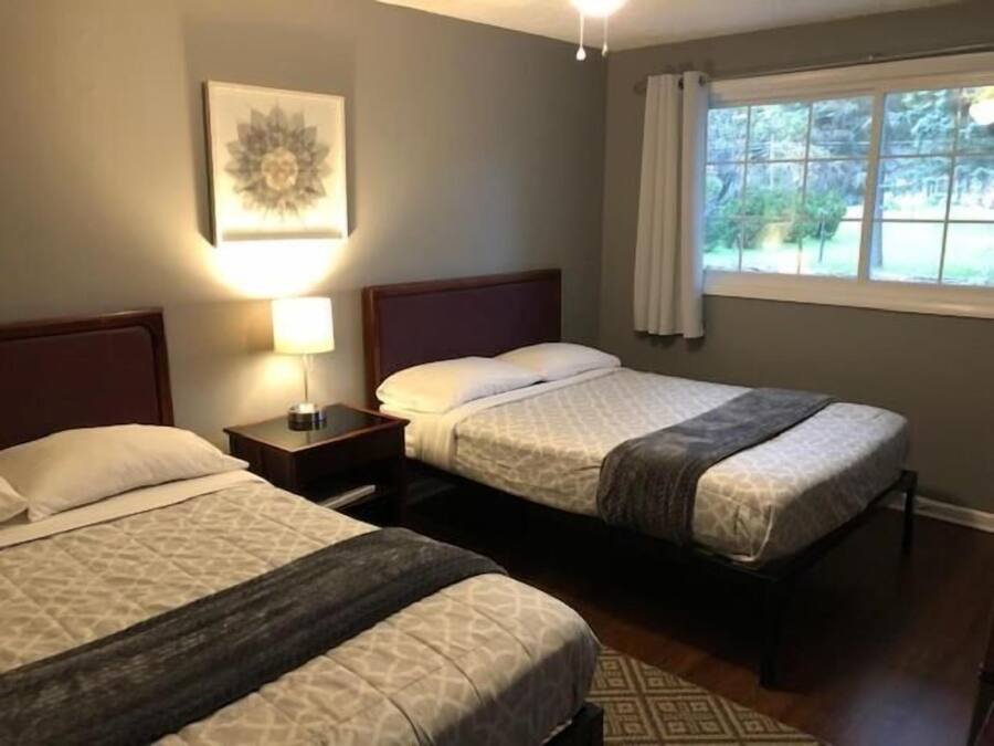 Habitación de hotel con dos camas, una mesita de noche con lámpara, un cuadro enmarcado en la pared y una ventana con cortinas.