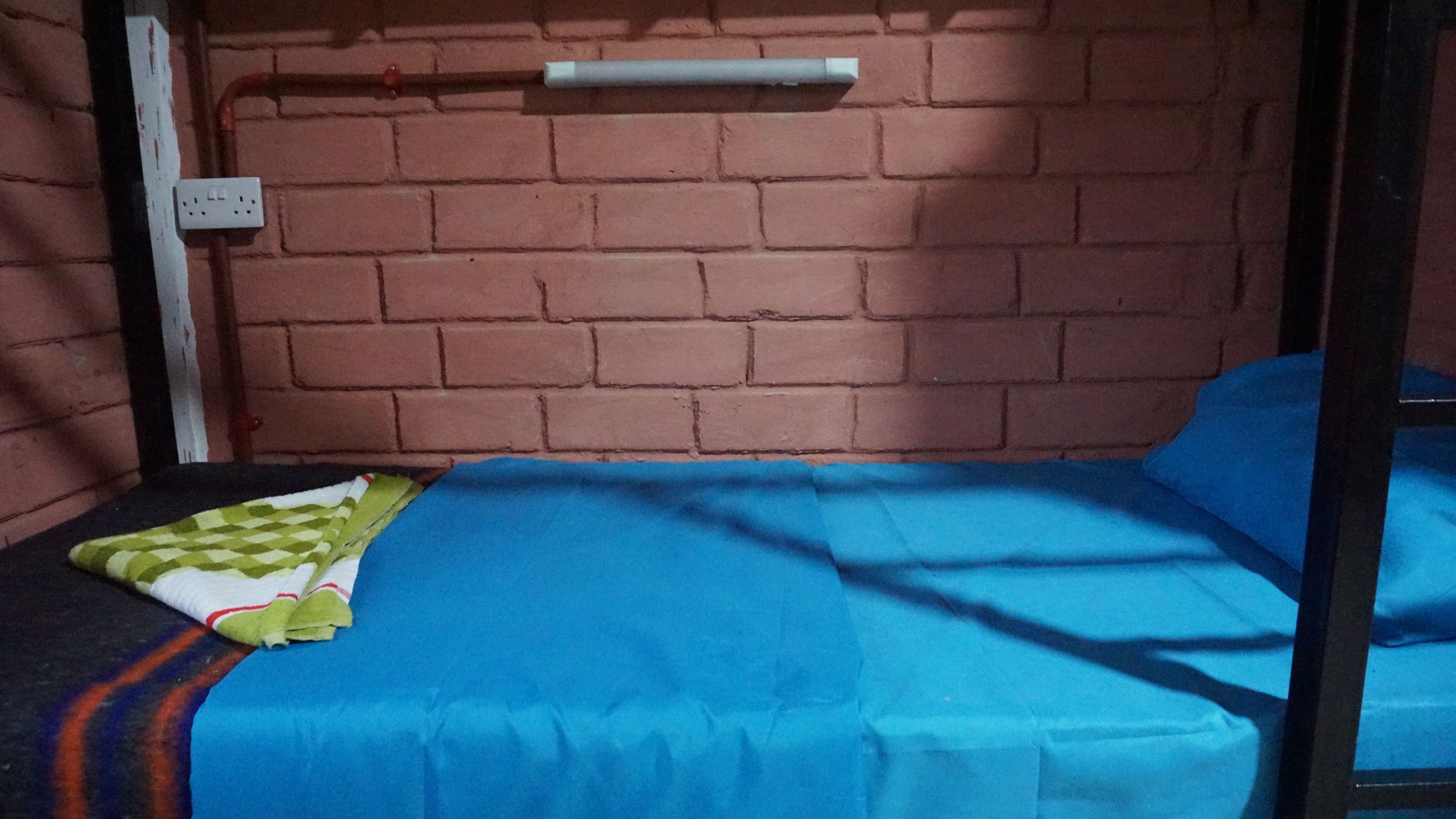 Un letto singolo con un materasso blu e una coperta bianco-verde. Si nota una presa elettrica bianca sulla parete in mattoni.