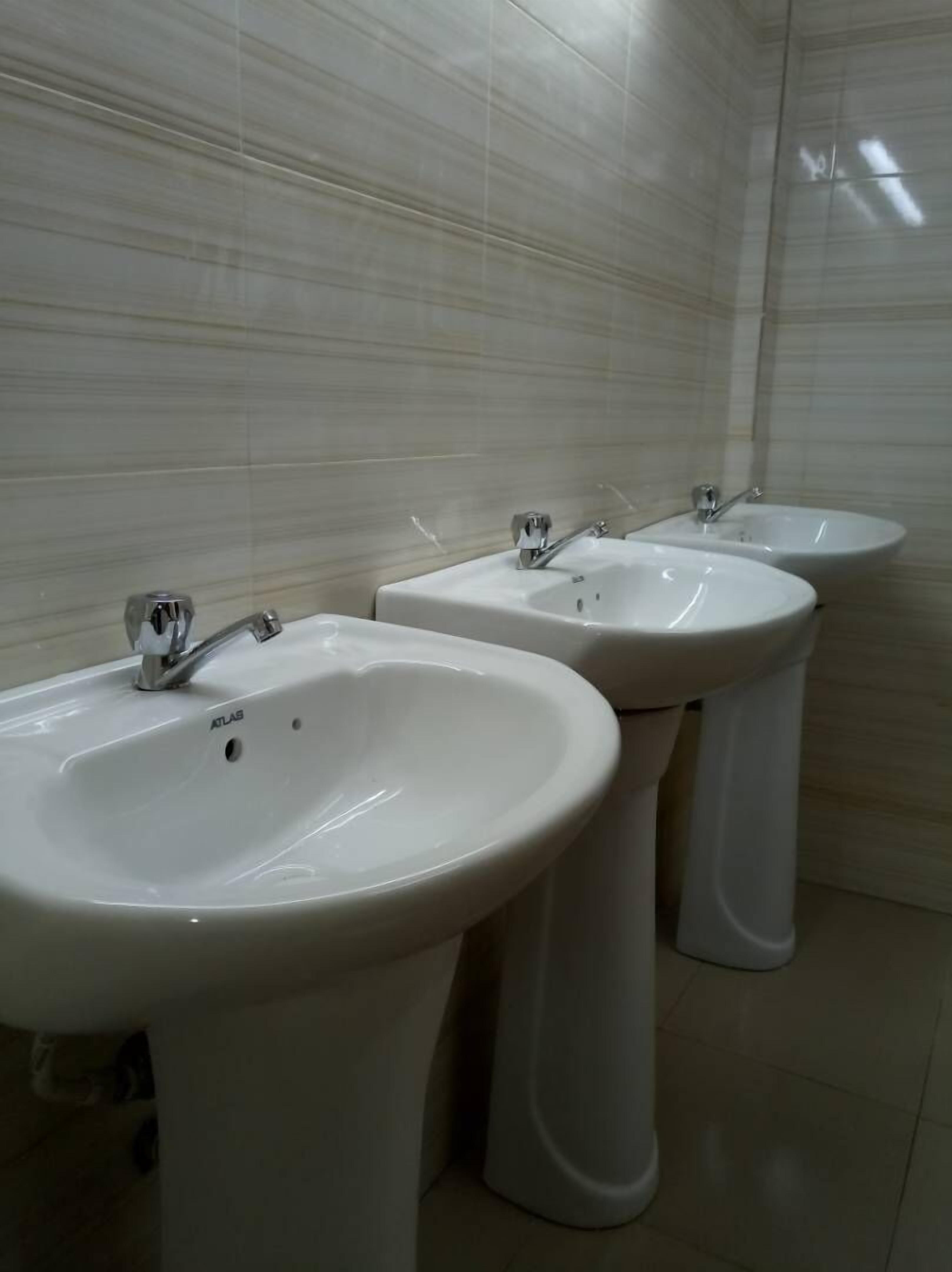 Three white pedestal sinks with faucets in a bathroom with tiled walls and floor.