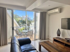 Una sala de estar moderna con un sofá de cuero, una mesa de centro de madera, un televisor y un balcón con vistas a palmeras y edificios.