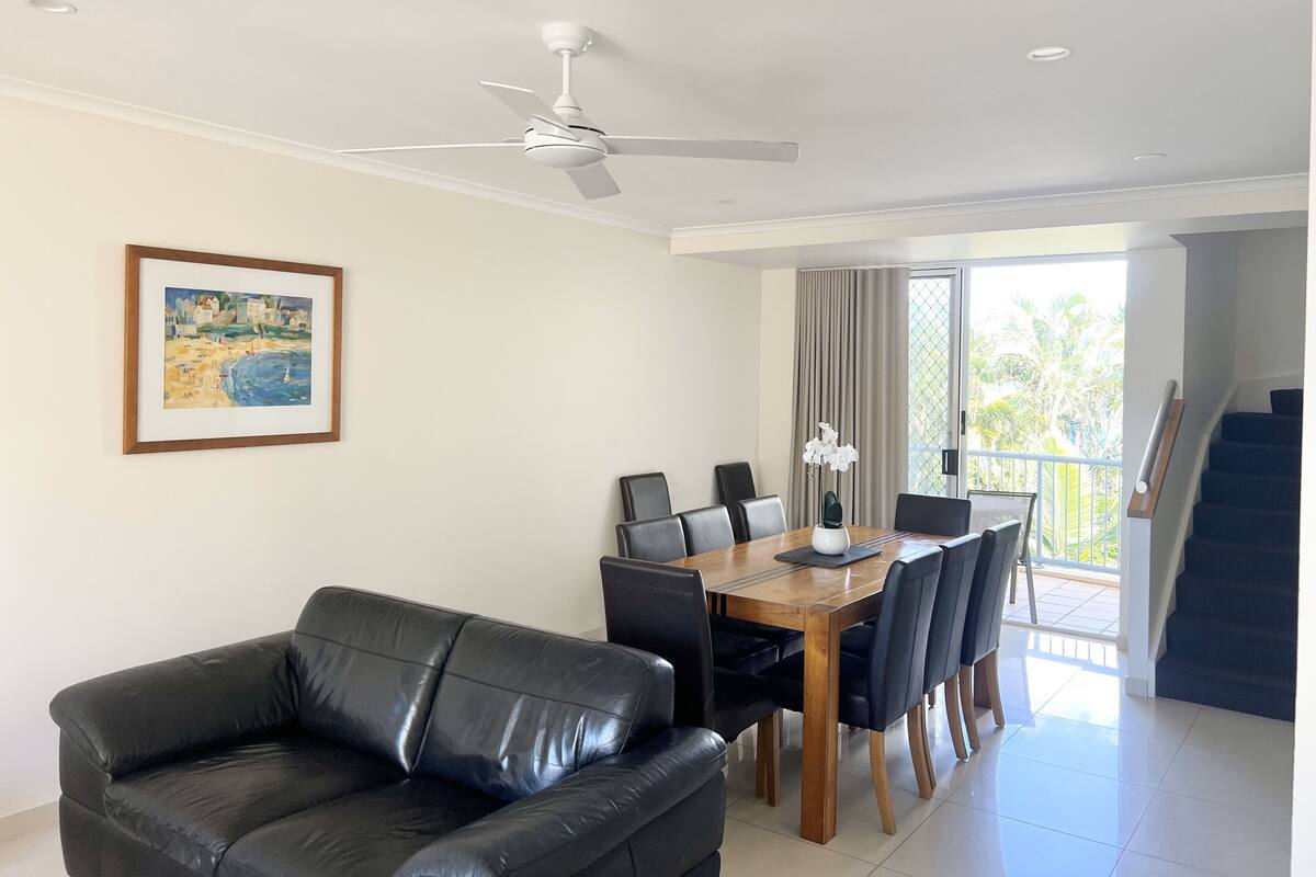 A dining area with a wooden table and chairs, a black leather sofa, a ceiling fan, and a framed painting on the wall.