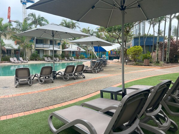 Un espace aménagé au bord de la piscine, avec des chaises longues et des parasols, entouré de palmiers et d’un bâtiment doté de balcons.