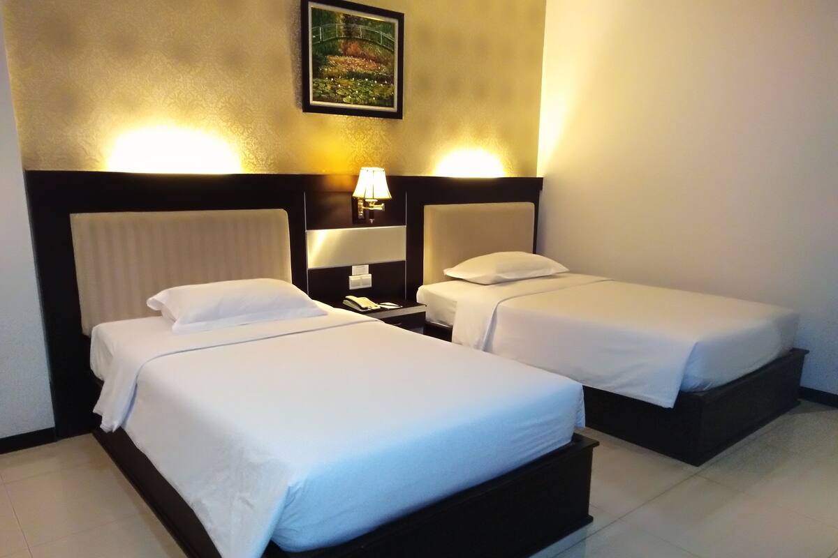 A hotel room with two single beds, a nightstand with a lamp, a painting on the wall, and a tiled floor.