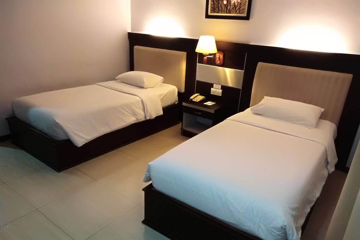 A hotel room with two single beds, a nightstand with a lamp, and a telephone.