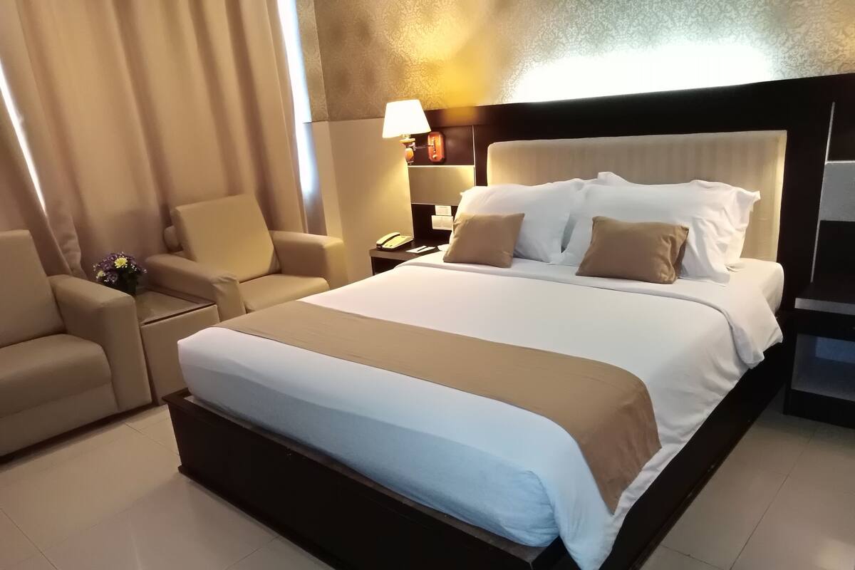 A hotel room with a large bed, a bedside table with a lamp, a chair, and a small table with a vase of flowers.