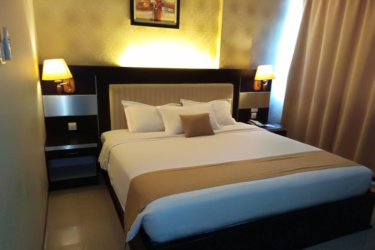 A neatly made bed with white linens and a brown throw, flanked by two bedside tables with lamps, in a hotel room with a plain wall and a window with curtains.