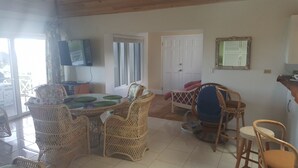 A dining area with wicker chairs and a glass-top table, a television mounted on the wall, and a door leading to another room. - Horizon House (Governor's Harbour)